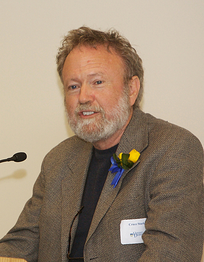 ortrait of a bearded man speaking at a microphone, wearing a textured blazer with a blue-and-yellow boutonniere pinned to the lapel. He appears seated at an event or panel, with a name tag visible on his jacket and a plain light background behind him.