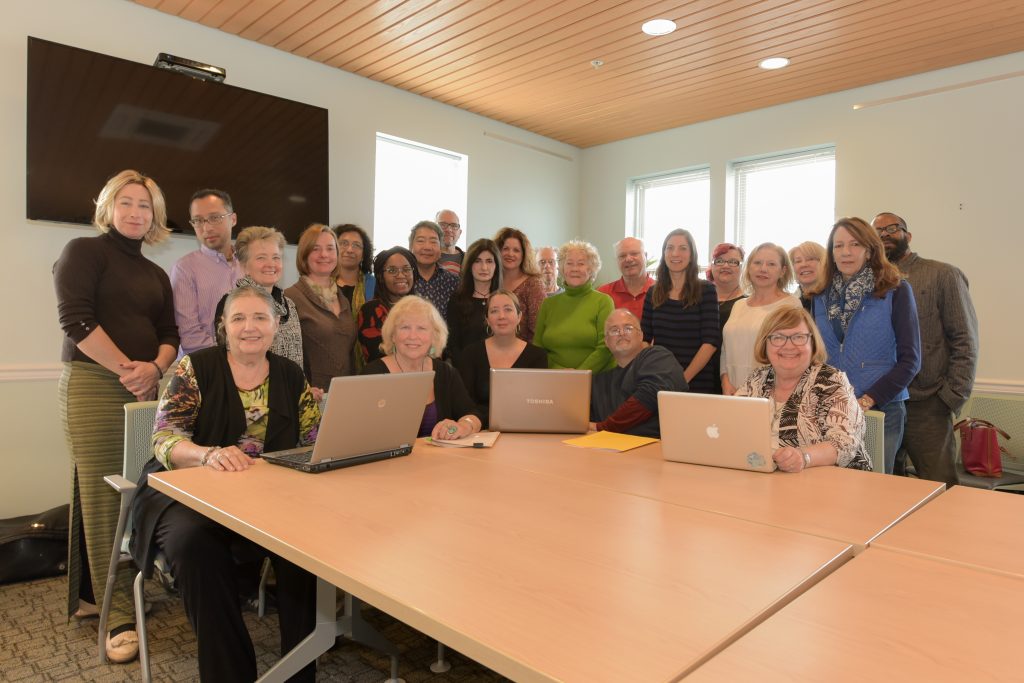 Large group photo of workshop participants gathered around a conference table in a bright meeting room. Several people sit at the table with open laptops while others stand behind them, smiling toward the camera beneath a wall-mounted screen and tall windows.