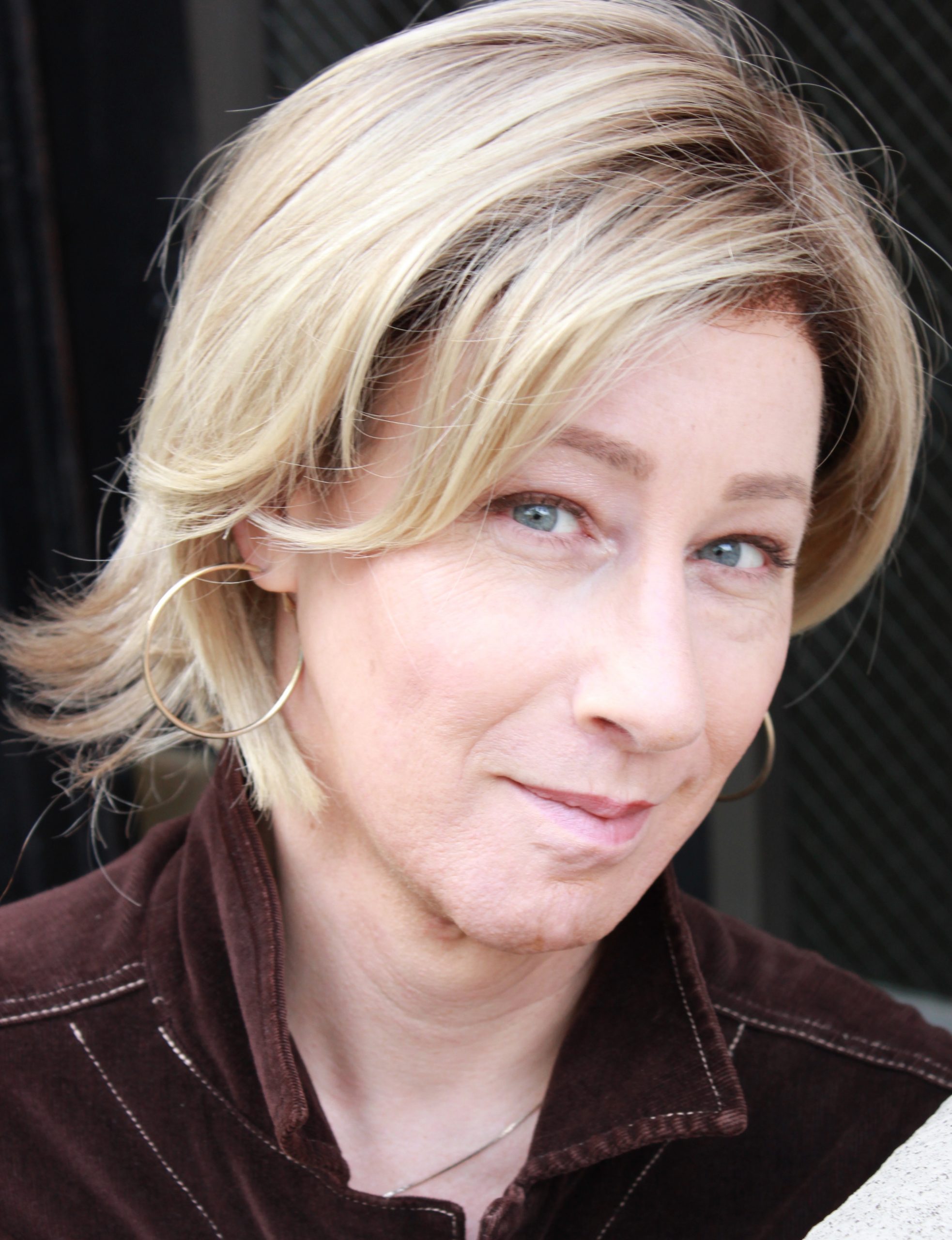 Close-up portrait of a woman with short blonde hair and hoop earrings, looking toward the camera with a composed expression. She wears a dark collared jacket, and the softly blurred background keeps the focus on her face.