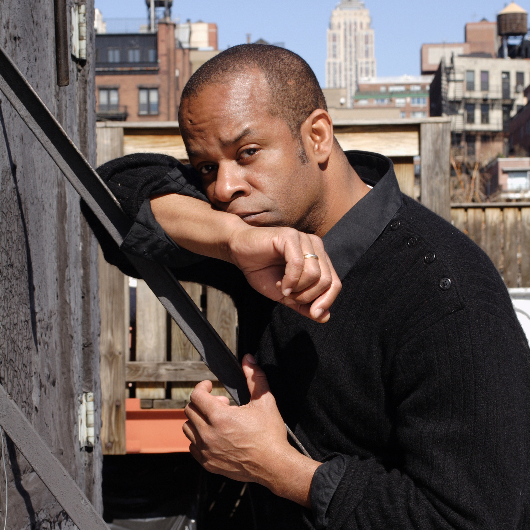 Portrait of a man leaning on an outdoor metal stair railing on a rooftop or terrace, with city buildings blurred in the background. He wears a dark sweater and rests his face on one arm, looking directly at the camera with a thoughtful, serious expression.