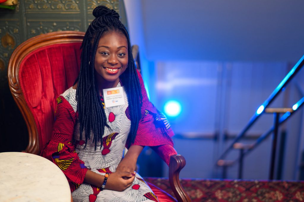 A smiling Poetry Out Loud participant with long braids sits in a red upholstered chair wearing a patterned dress and a name badge, with soft blue stage lighting and stairs in the background.