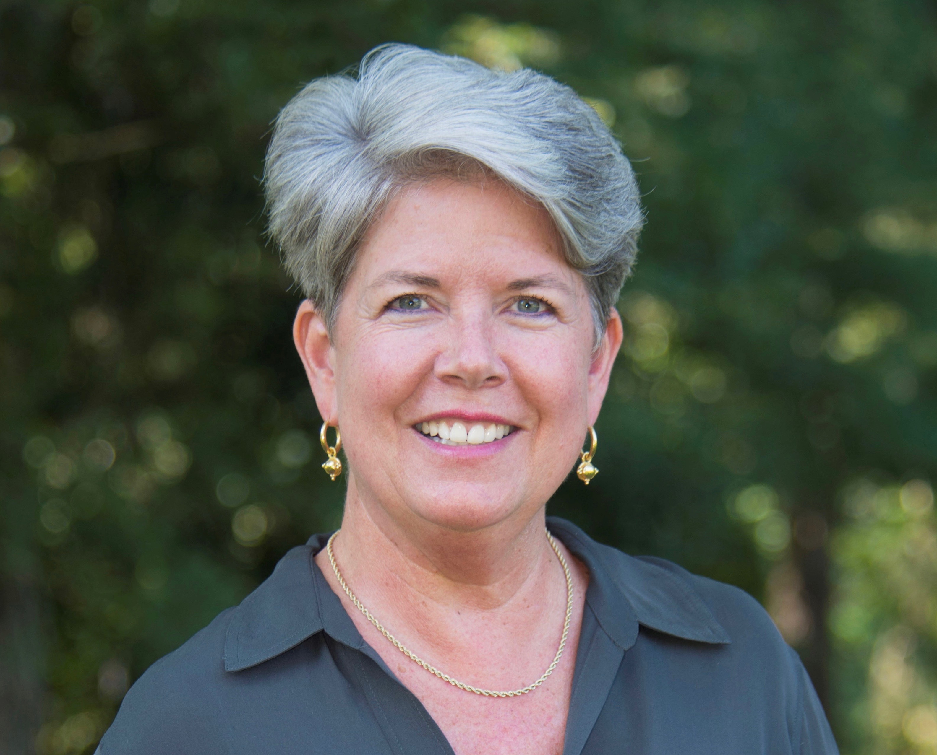 A smiling woman wearing a black shirt.  She is outside and behind her are trees.