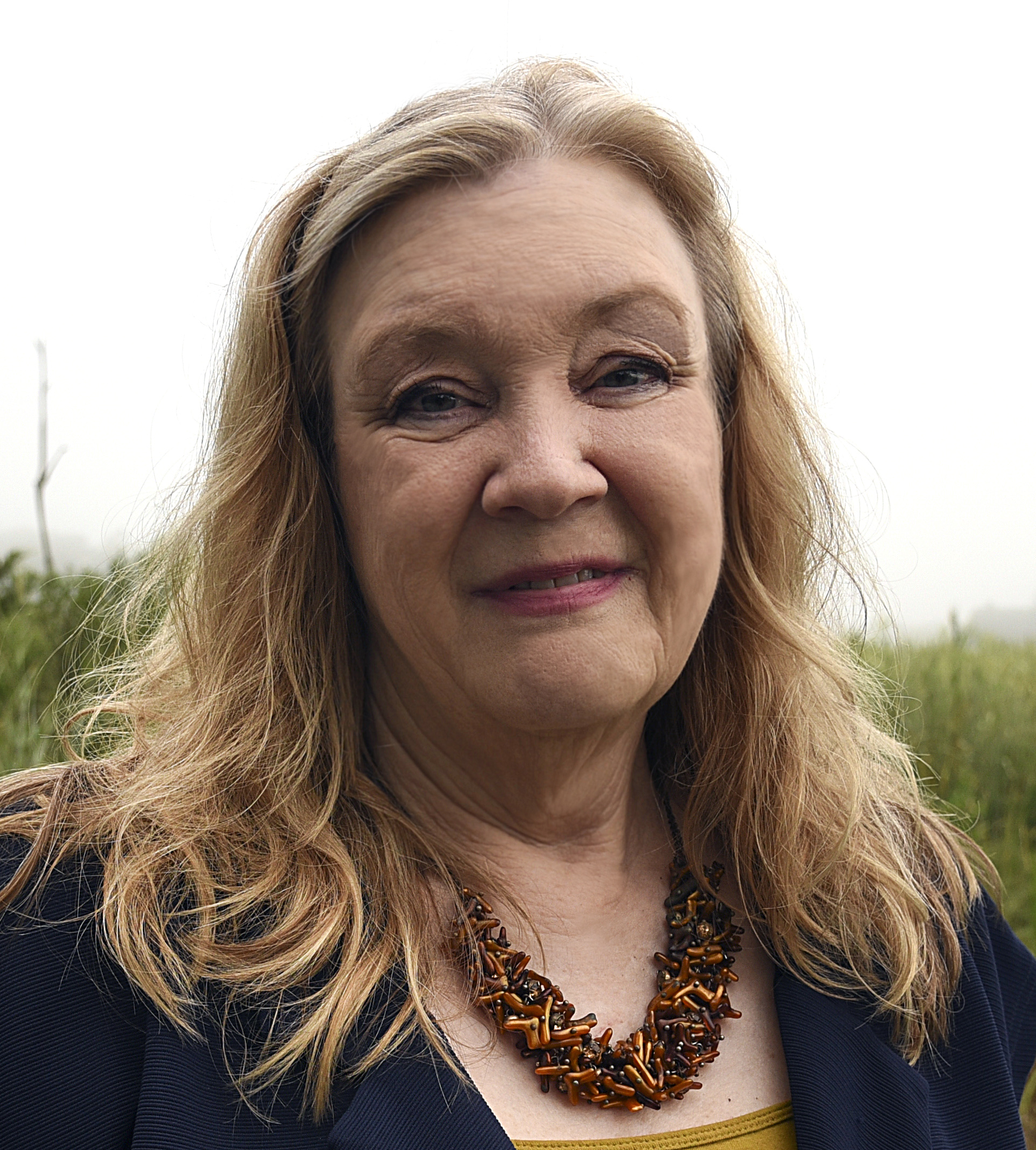 A woman with blonde hair, black shirt, and necklace smiling.  She is outside with greenery behind her.