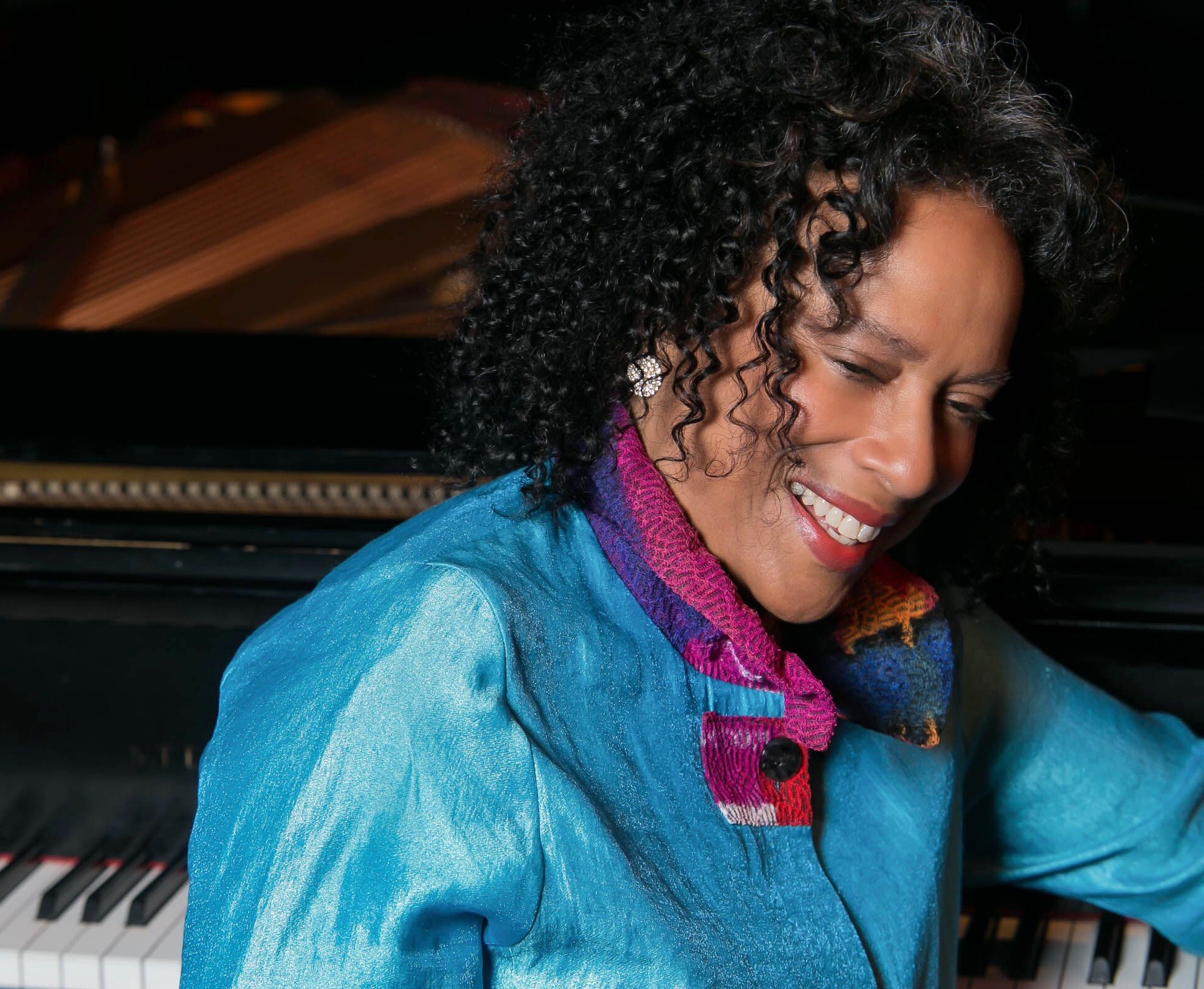 A woman wearing a blue shirt and multi colored scarf seated at a piano and smiling.
