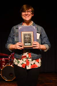 three quarter body portrait of Camille Decker 2020 Poetry Out Loud Delaware State Champion holding her plaque and certificate in front of a dark background and drumset