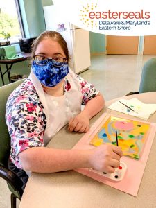Woman lookinWomaA woman is looking at the camera while holding a paintbrush, pausing her painting for the picture. She is sitting at a table in an Easterseals facility center.