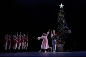 Spotlight on the female dancer in pink dress who stands with arms outreached next to a male dancer in a soldier's uniform holding a sword in front of a Christmas tree with presents beneath it on an otherwise dark stage; a man in a dark cloak also stands in front of the Christmas tree but more in the background and two rows of dancers dressed in a different military uniform stand to the left of the Christmas tree scene