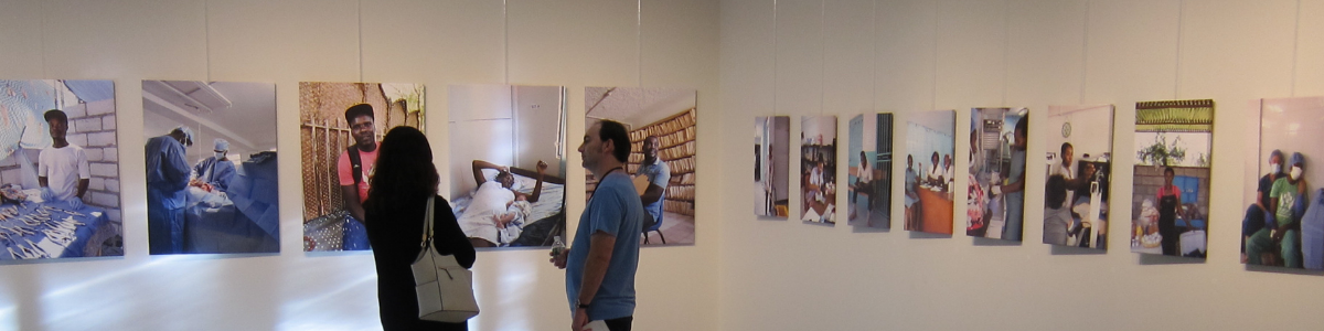 A woman and a man are standing in the Mezzanine Gallery, looking at the pictures on exhibit. The woman's back is towards the camera, while the man is standing to face the woman and look at the pictures at the same time. The pictures are hanging on two white walls that meet at a corner.