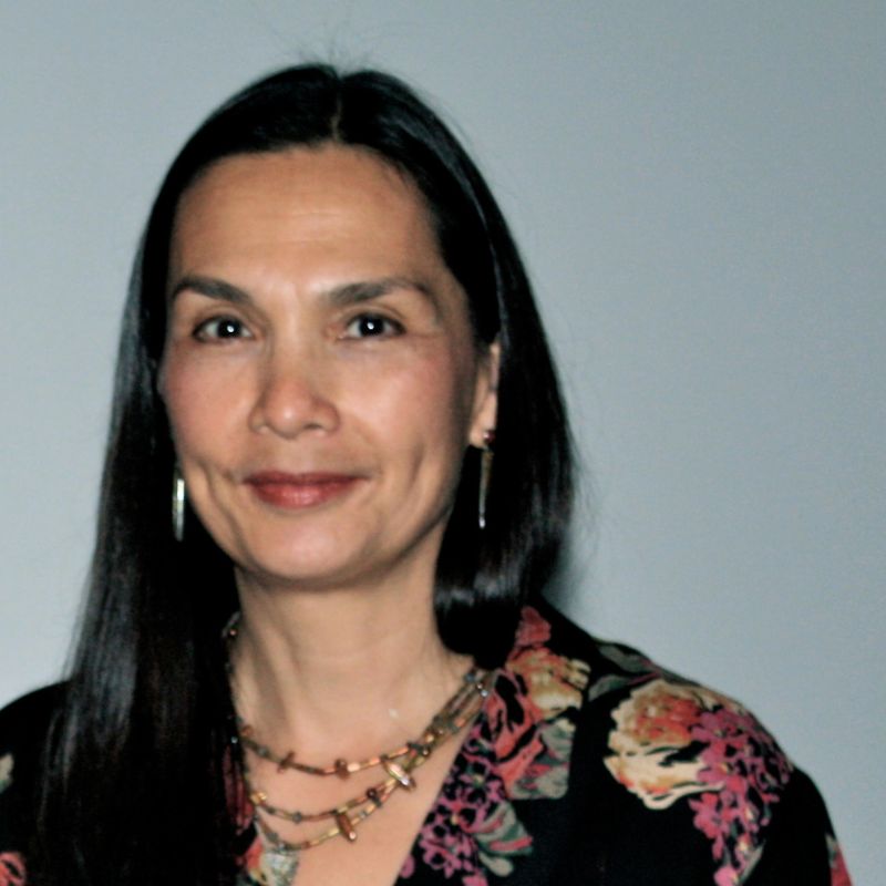 Headshot of a woman with long dark hair wearing a black floral blouse, layered necklaces, and earrings. She smiles gently at the camera against a plain light gray background.