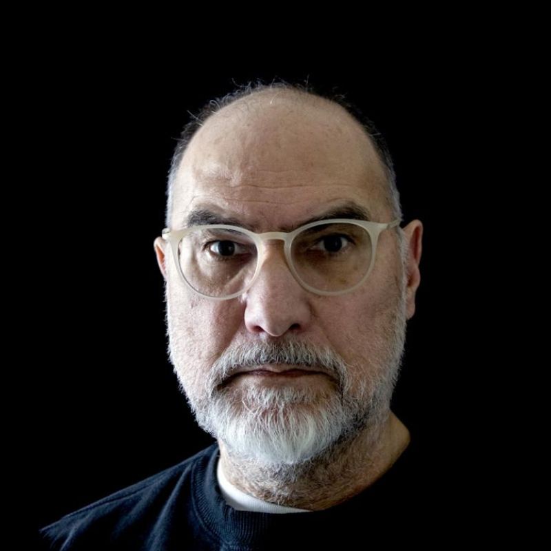 A headshot of a bald white man with a gray beard and round glasses, in front of a black background.