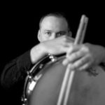 A headshot of a white man wearing a collared shirt, in front of a black background with drumsticks
