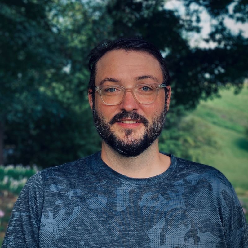 A man with dark hair, a mustache and a beard, in a blue shirt and glasses, standing outside.
