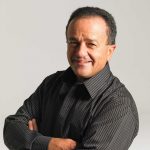 A man in a dark gray button up shirt, crossing his arms and smiling, in front of a light gray background.