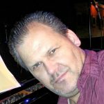 A white man in a purple shirt, in front of keyboards.