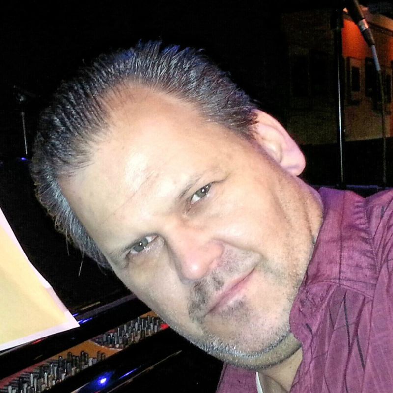 A white man in a purple shirt, in front of keyboards.