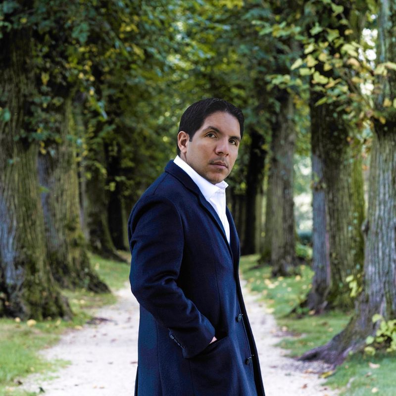 A man standing between rows of trees. He is wearing a blue suit.
