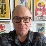 A headshot of a white man in glasses and a gray flannel shirt, in front of a gallery wall.
