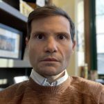 A headshot of a white man with a collared shit under a brown sweater, inside.