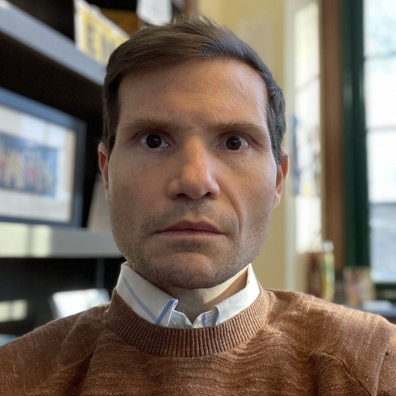 A headshot of a white man with a collared shit under a brown sweater, inside.