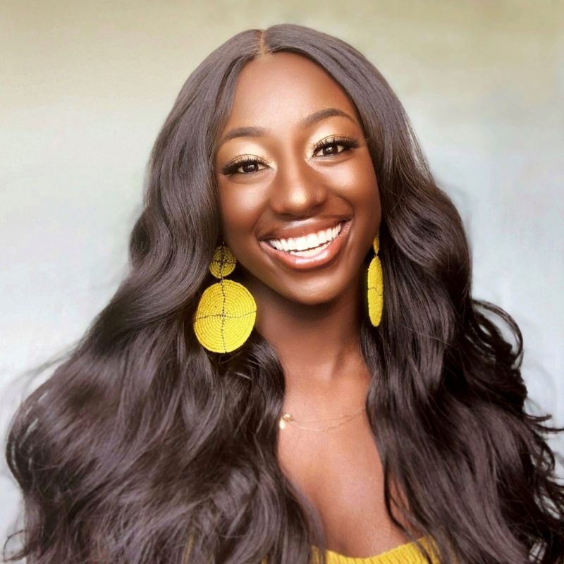 A headshot of a young black woman with long hair, wearing yellow earrings and a yellow dress, smiling.
