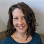 A picture of a white woman with dark hair in a blue shirt, sitting in front of a white wall.