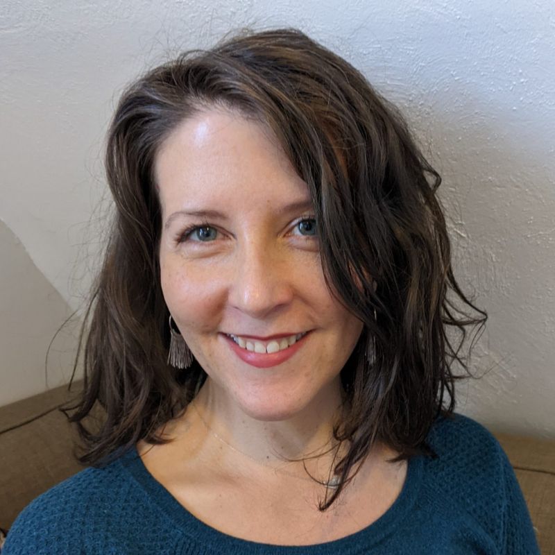 A picture of a white woman with dark hair in a blue shirt, sitting in front of a white wall.