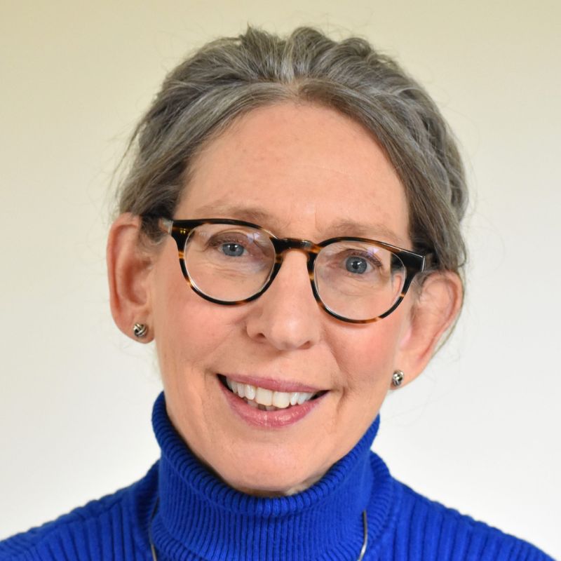 A headshot of a white woman in a blue turtleneck sweater and glasses, in front of a white background.