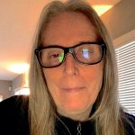A white woman in black glasses and a black sweater, with medium length blonde hair.