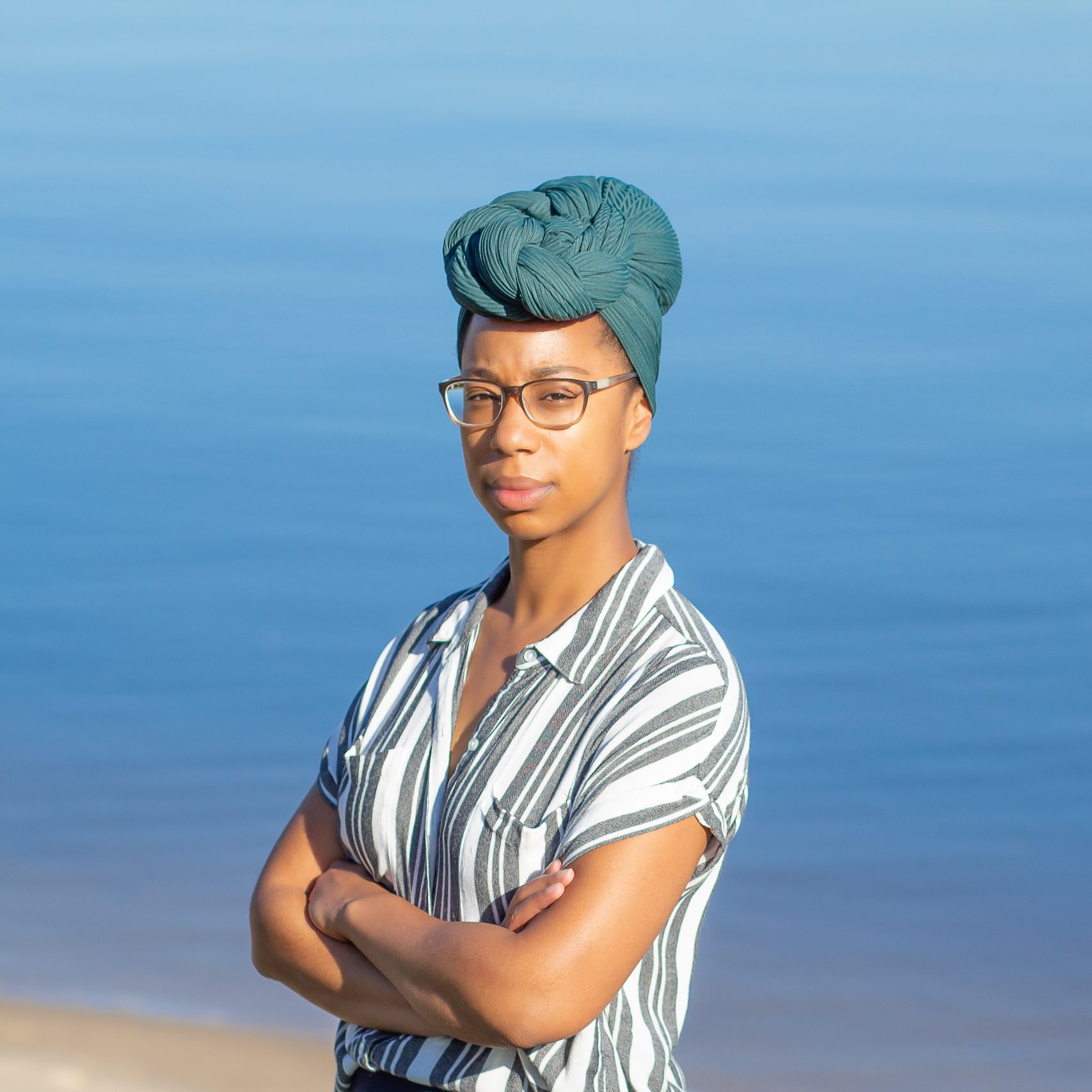 Portrait of a person standing outdoors near calm blue water, arms crossed and looking at the camera. They wear glasses, a green headwrap, and a striped short-sleeved shirt, with the soft shoreline and water creating a simple, serene background.