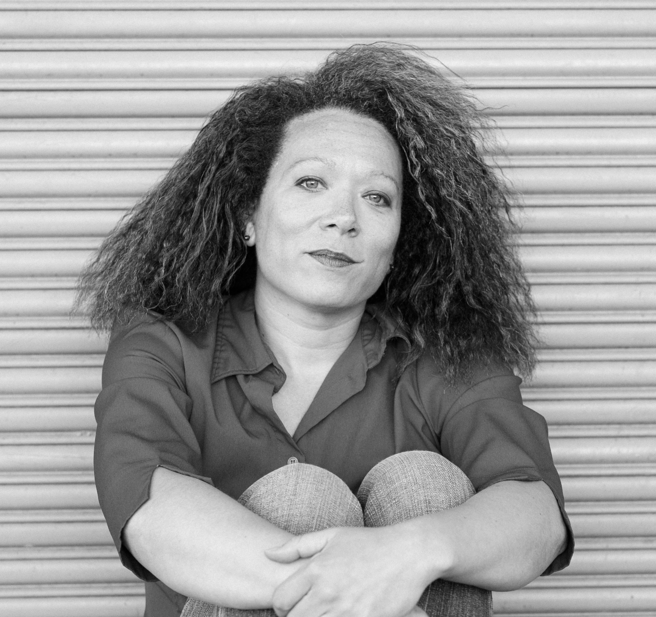 Black-and-white portrait of a woman seated with her arms wrapped around her knees in front of a metal roll-up door. She looks toward the camera with a calm, reflective expression, and her voluminous curly hair frames her face.