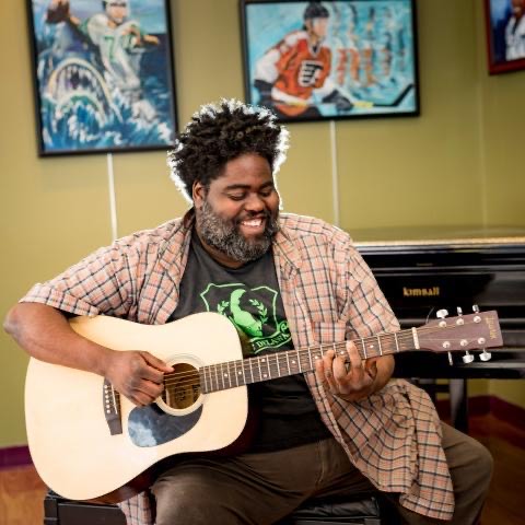A man holding a guitar with black hair and a beard. He is sitting in front of a green wall with paintings on it.
