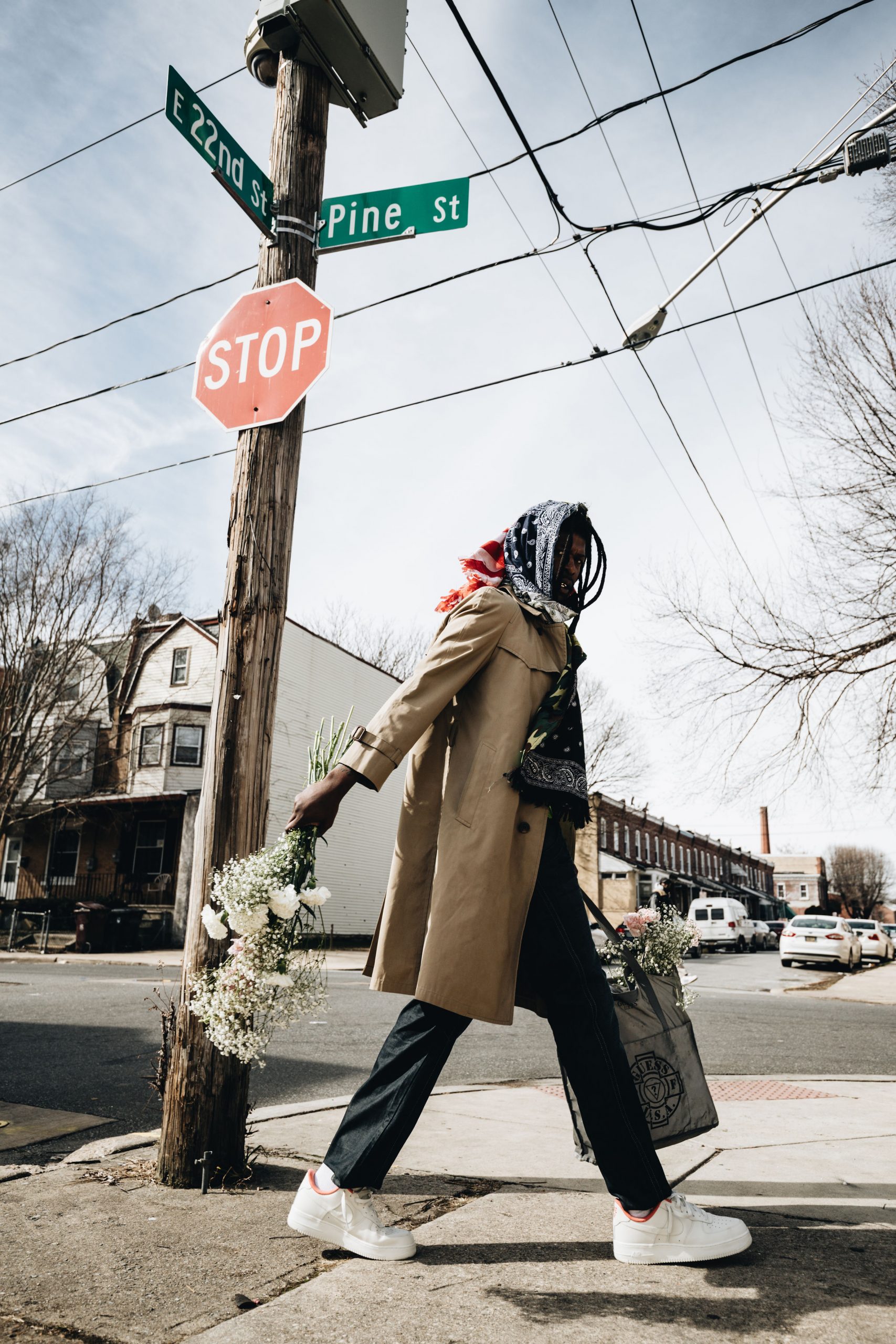 A photograph of a person walking in a coat and a headscarf, past a stop sign in a neighborhood. They are holding a bag of white flowers.