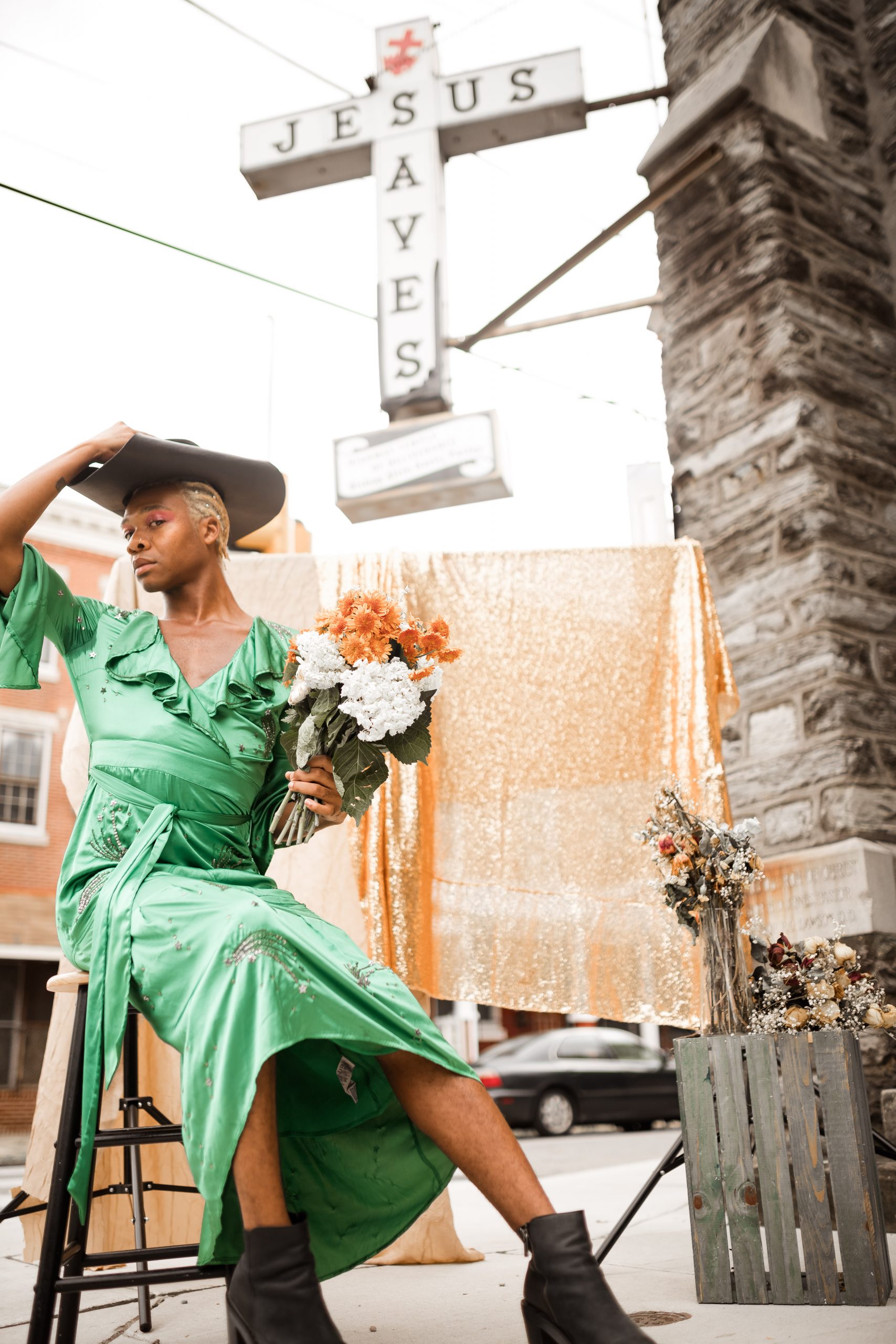 A photograph of a black person sitting on a stool in a green dress and a hat, outside of a church.