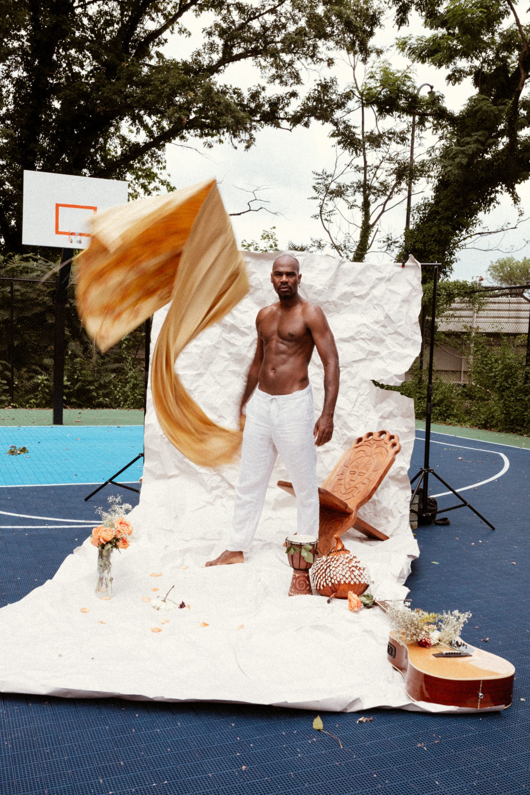 A black man shirtless in white pants on a white backdrop in the middle of a basketball court, surrounded by brown, orange, and gold props.