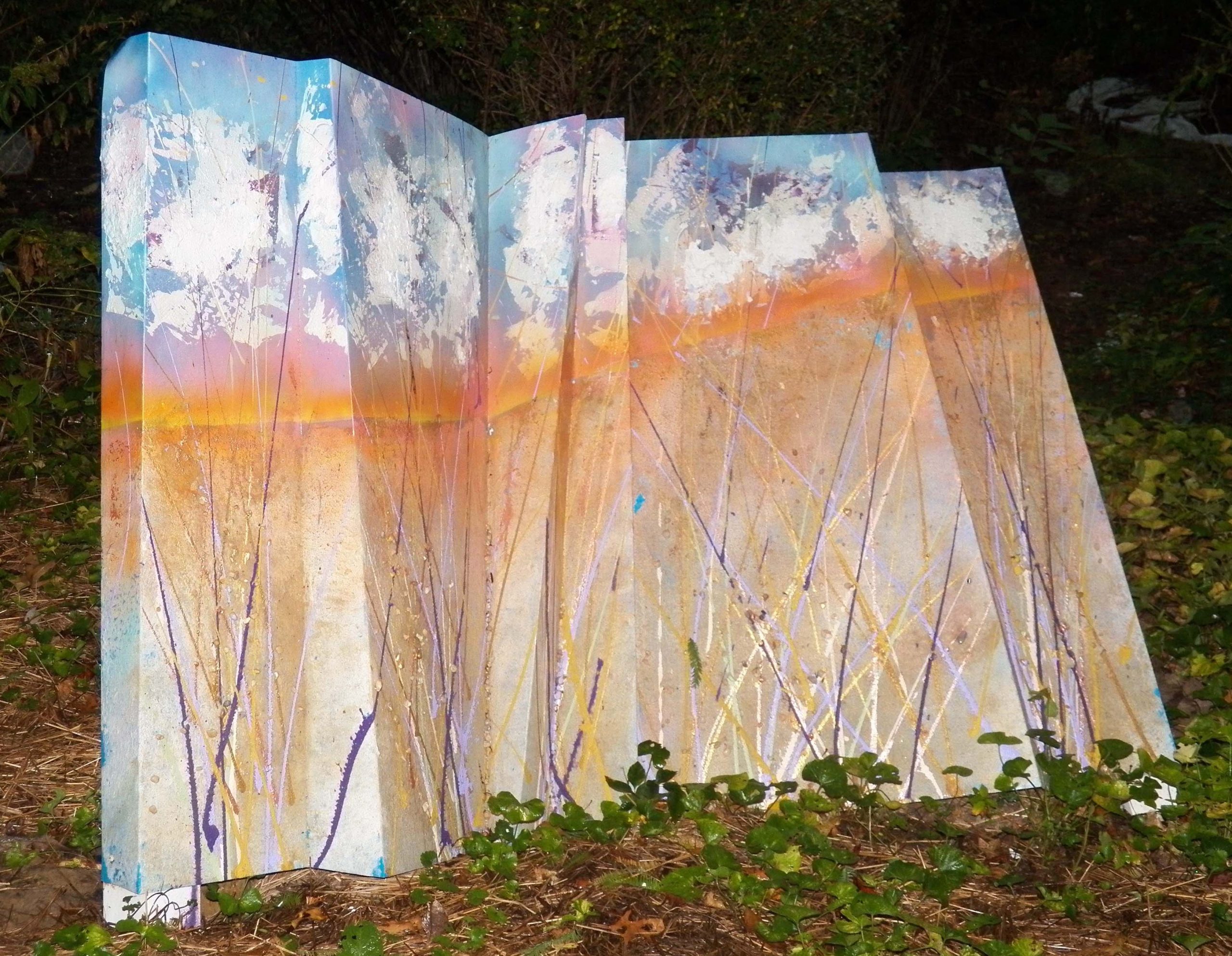 A large, painted aluminum slab standing on grass. It is painted to look like a daytime landscape of dry fields.