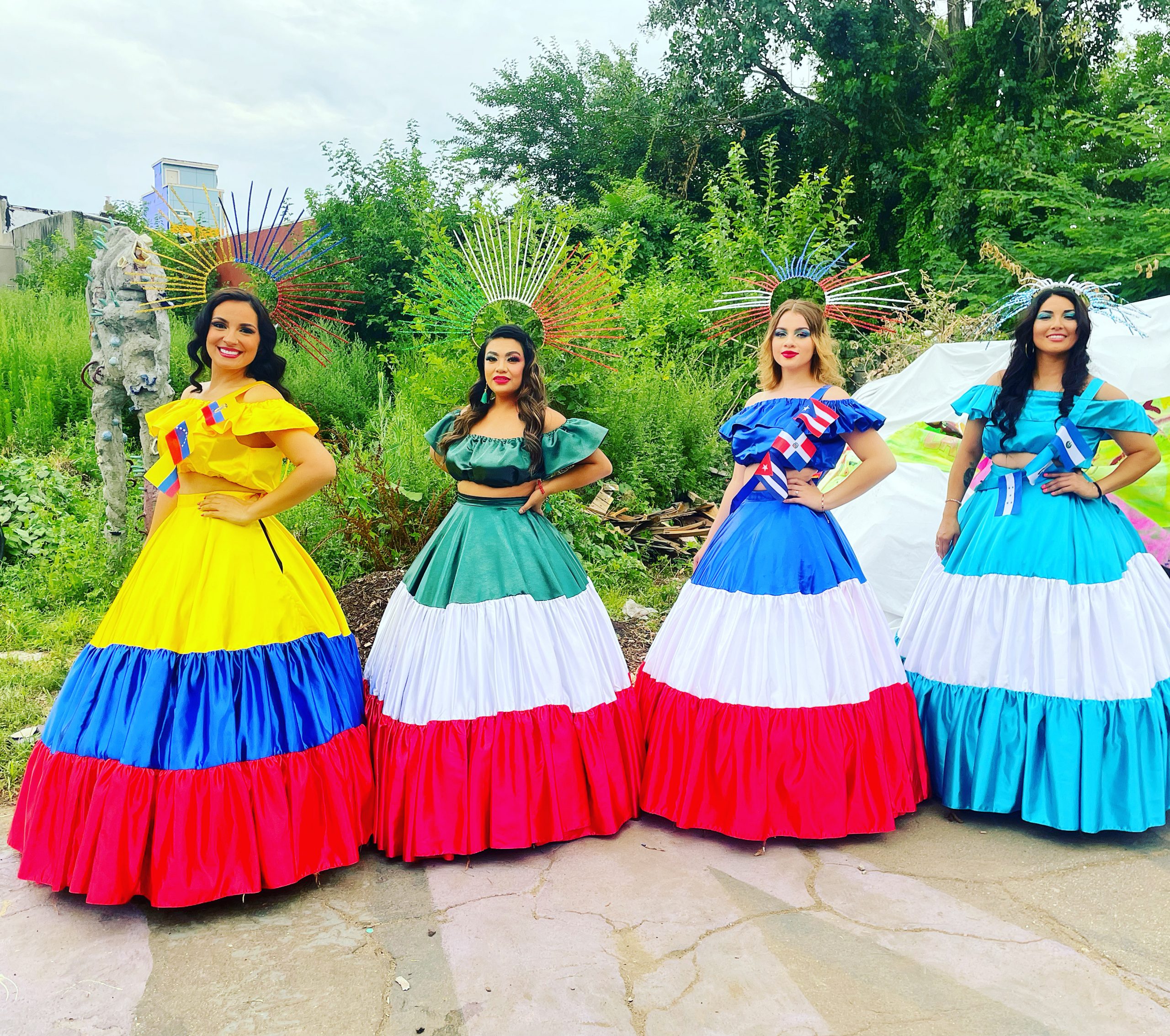 Four women wearing large dresses with the colors of different countries, seen outside.