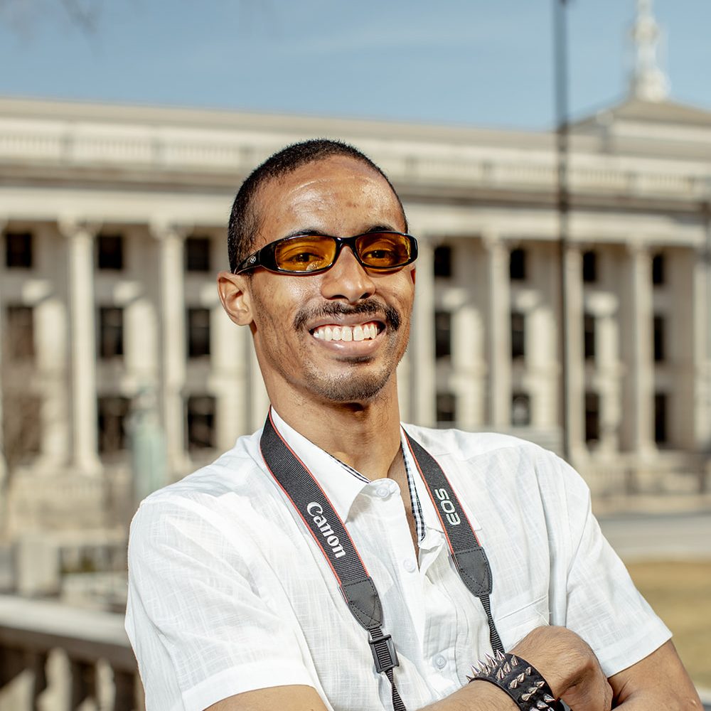 A black man with a mustache wearing a short-sleeved button-up shirt, with a camera around his neck, outside.
