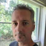 A white man with a goatee in front of a window during the day.