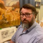 A white man with a beard, wearing a button up shirt, in front of some paintings.