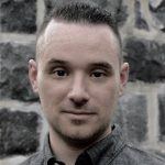 A white man's headshot in front of a stone brick wall.