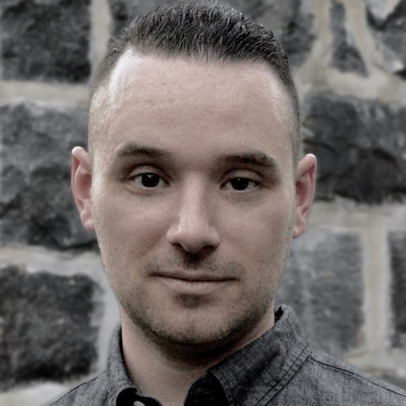A white man's headshot in front of a stone brick wall.