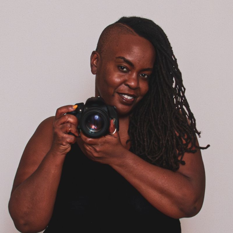 A black woman with a side-shave haircut, wearing a black tank tip and holding a camera.