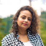 An Indian woman with curly hair, wearing a black and white flannel shirt, outside.