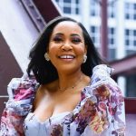 A black woman in a white floral dress, smiling, outside in front of a building.