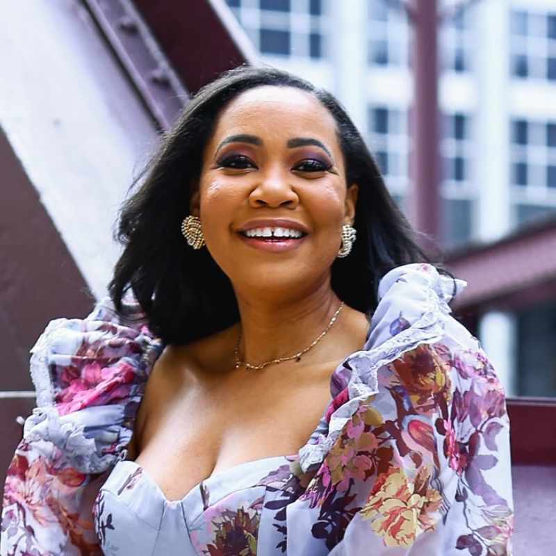 A black woman in a white floral dress, smiling, outside in front of a building.