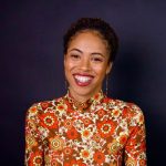 A black woman wearing an orange, floral, long-sleeve shirt. She is smiling in front of a dark blue wall.