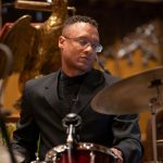 a man in a black suit with glasses playing the drums