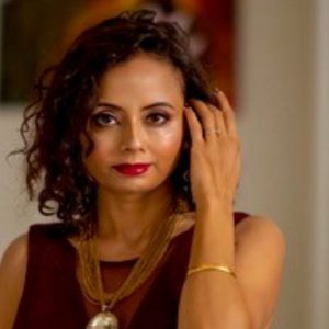 A woman with curly dark hair, wearing a sleeveless burgundy top, layered gold necklaces with a large pendant, and a gold bracelet. She has bold makeup with red lipstick and is touching her hair while looking slightly to the side.