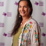 A woman with long dark hair wearing a green top and a white embroidered floral blouse, smiling in front of a step-and-repeat banner with the Texas Public Radio logo in purple.