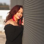 A woman with long, wavy red hair wearing a black off-the-shoulder dress, bright red tassel earrings, and a matching ring. She smiles while leaning against a textured metal wall in warm sunlight.
